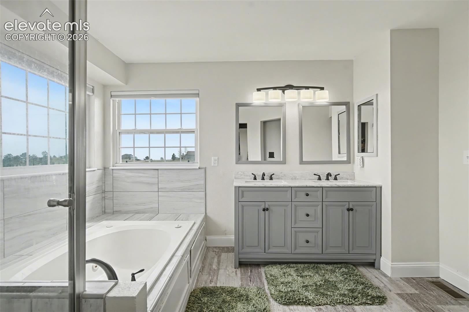 Dual-sink vanity with grey cabinetry and light-toned countertop, soaking tub with tile surround, wood-finish flooring, and multi-light fixture