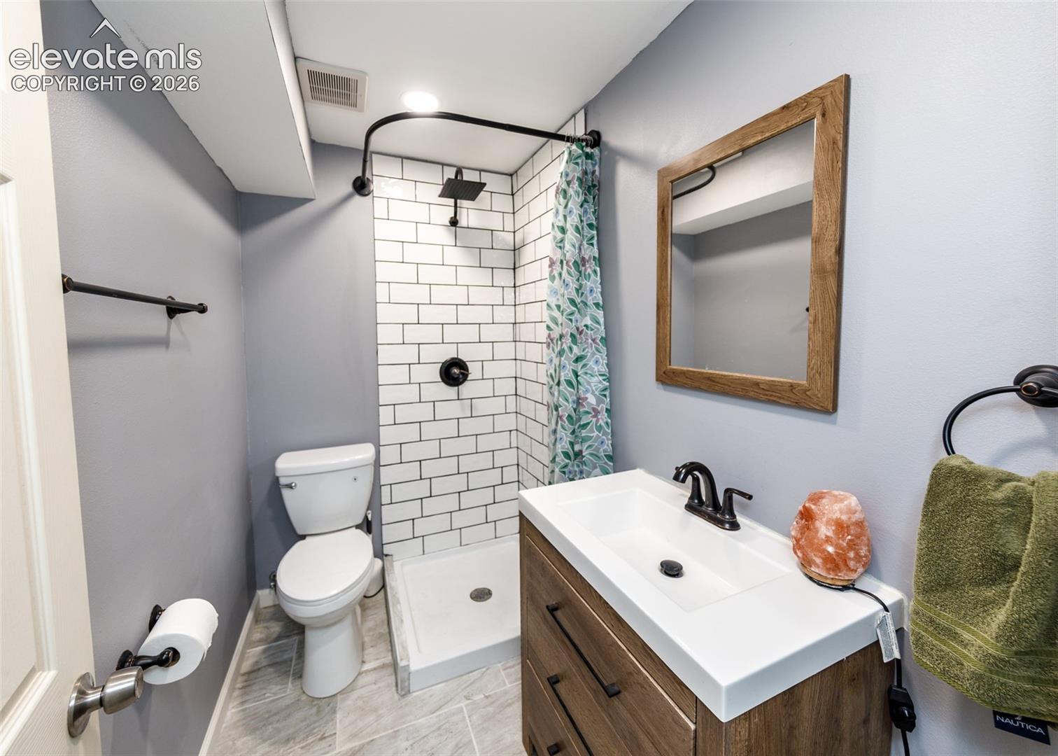 Modern bathroom featuring white subway tile shower surround with oil-rubbed bronze fixtures, wood-finish vanity with rectangular white sink, matte black faucet, framed mirror, and ceramic tile flooring