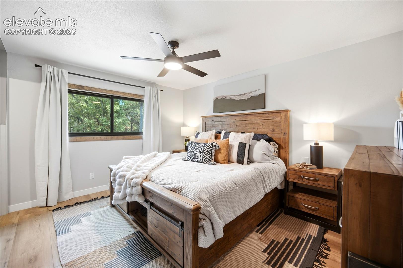 Neutral-toned bedroom featuring wood-finish flooring, a wood-trimmed window casing, and a multi-blade ceiling fan with integrated lighting