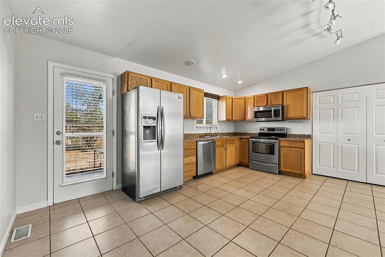 Kitchen featuring medium-toned wood cabinetry, stone-surface countertops, stainless steel appliances, ceramic tile flooring, and a glass-paneled exterior door