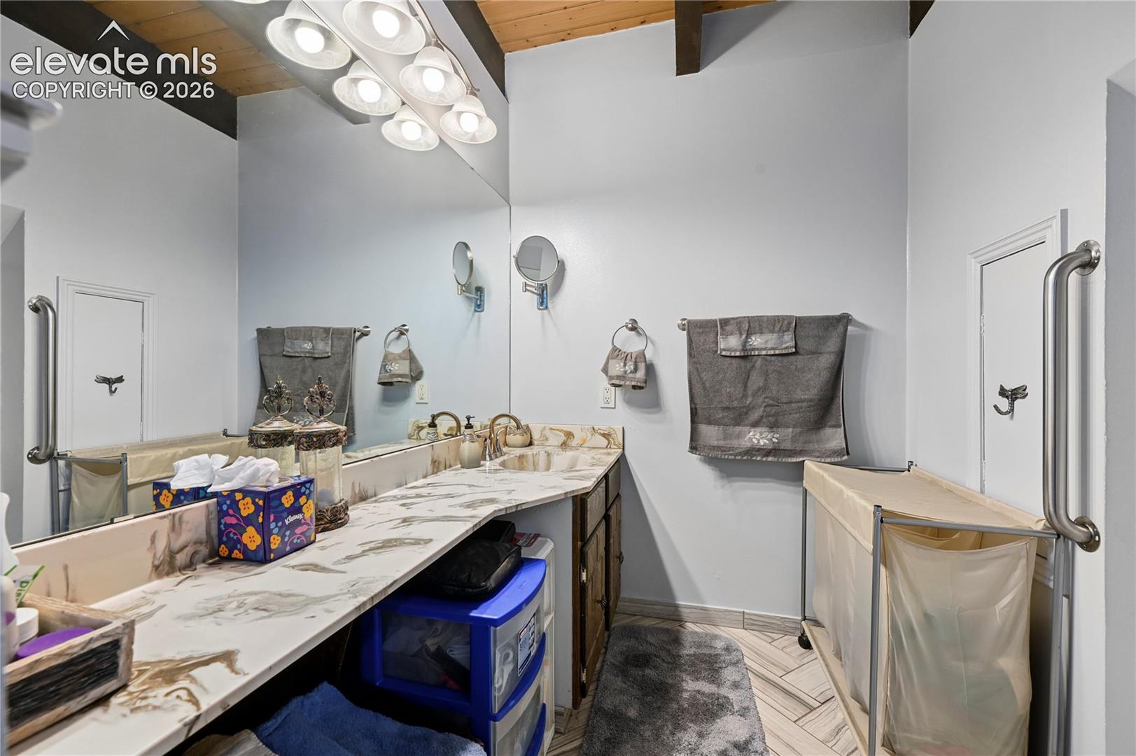 Main bathroom Dual vanity with integrated sinks, expansive wall-mounted mirror, overhead task lighting, and vaulted wood-paneled ceiling