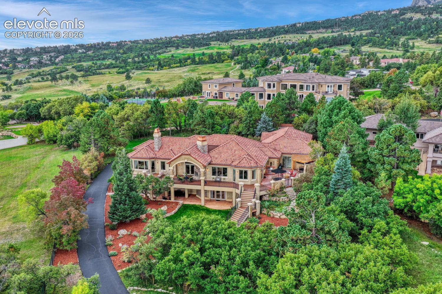 Multi-level residence featuring a clay tile roof, stone masonry chimneys, an elevated wraparound deck, and a paved driveway, set against a mountainous landscape