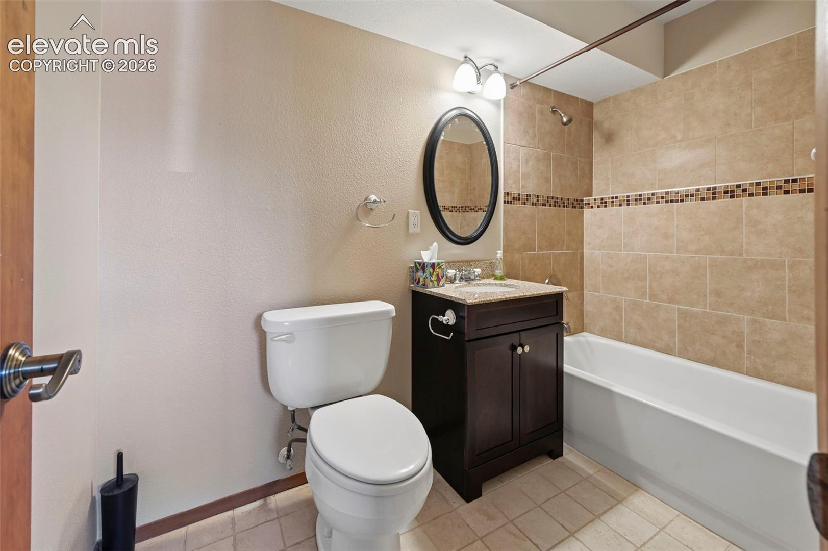 Dark-finish wood vanity with a stone countertop, oval-framed mirror, two-light fixture, and tiled tub surround featuring a decorative mosaic inlay