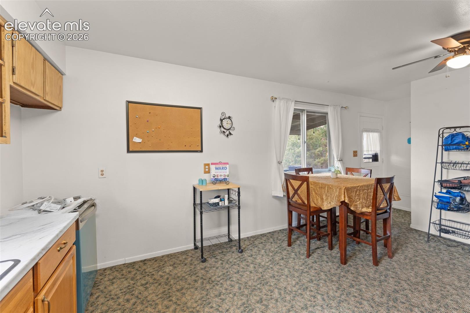 Wood cabinetry with gold-toned hardware, light-toned countertops, window with white curtains, patterned flooring, and a ceiling fan
