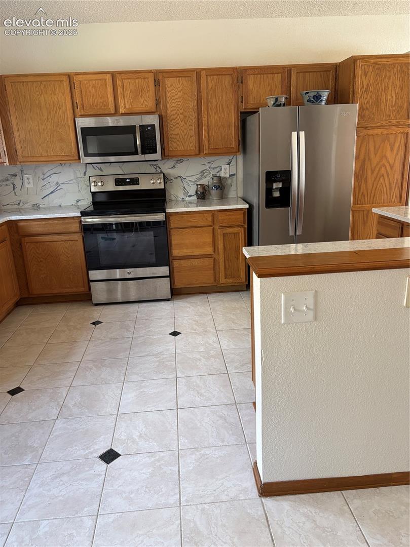 Kitchen featuring wood cabinetry, stone-finish countertops with a stone-patterned backsplash, stainless steel appliances, and tile flooring with decorative inserts