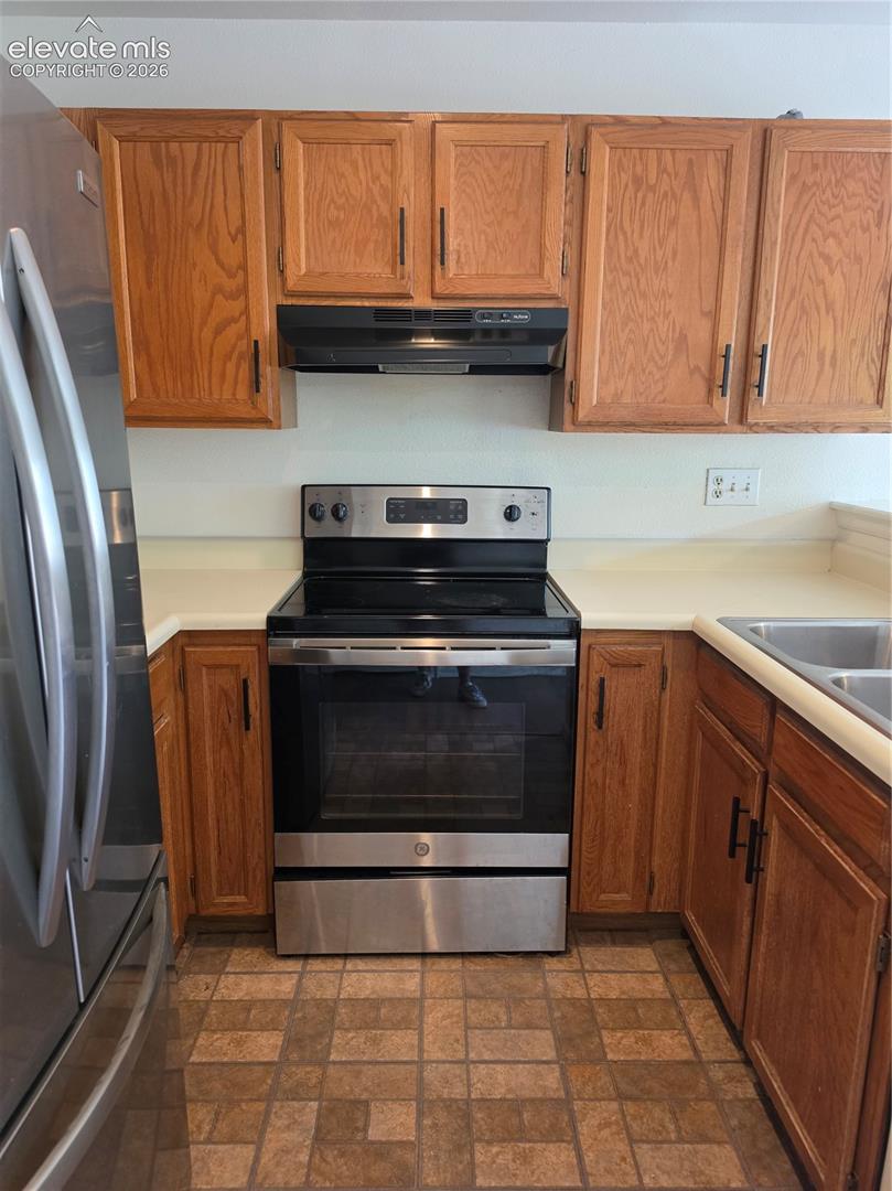 Kitchen featuring natural wood cabinetry, stainless steel appliances, solid-surface countertops, and ceramic tile flooring
