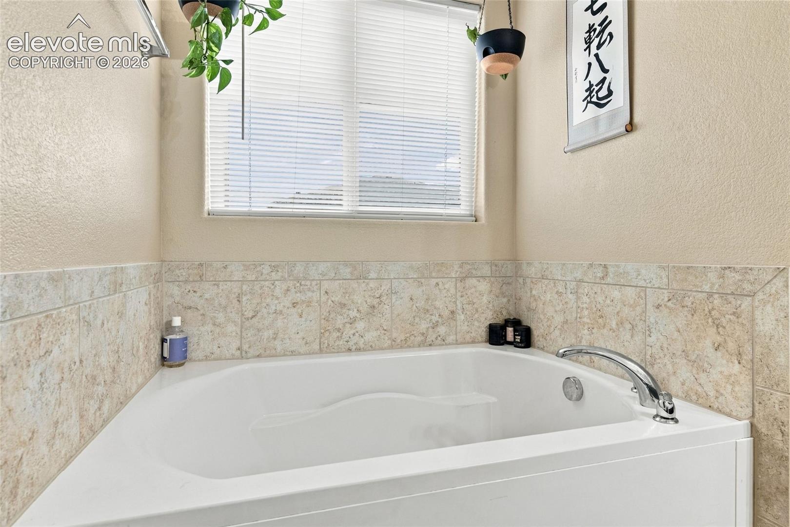 Built-in soaking tub with a chrome faucet, surrounded by neutral-toned tile wainscoting and a large window with horizontal blinds