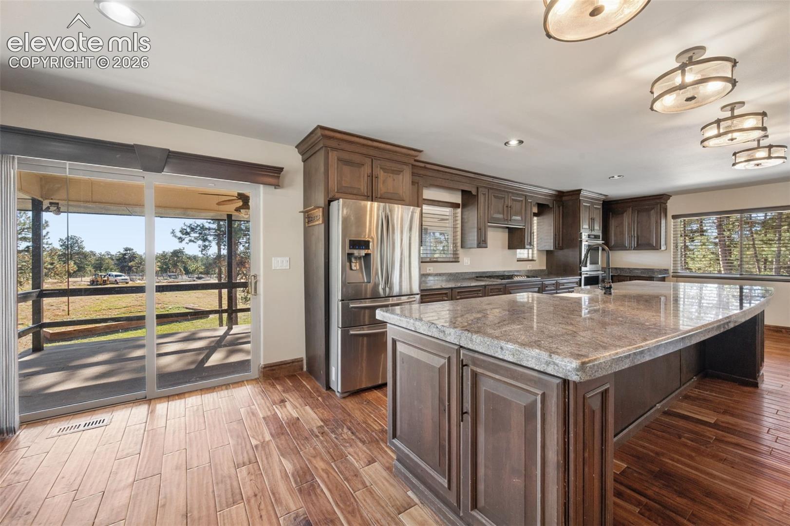 Kitchen featuring a large central island with stone countertops, dark-toned cabinetry, wood-finish flooring, stainless steel appliances, and sliding glass doors to the exterior deck