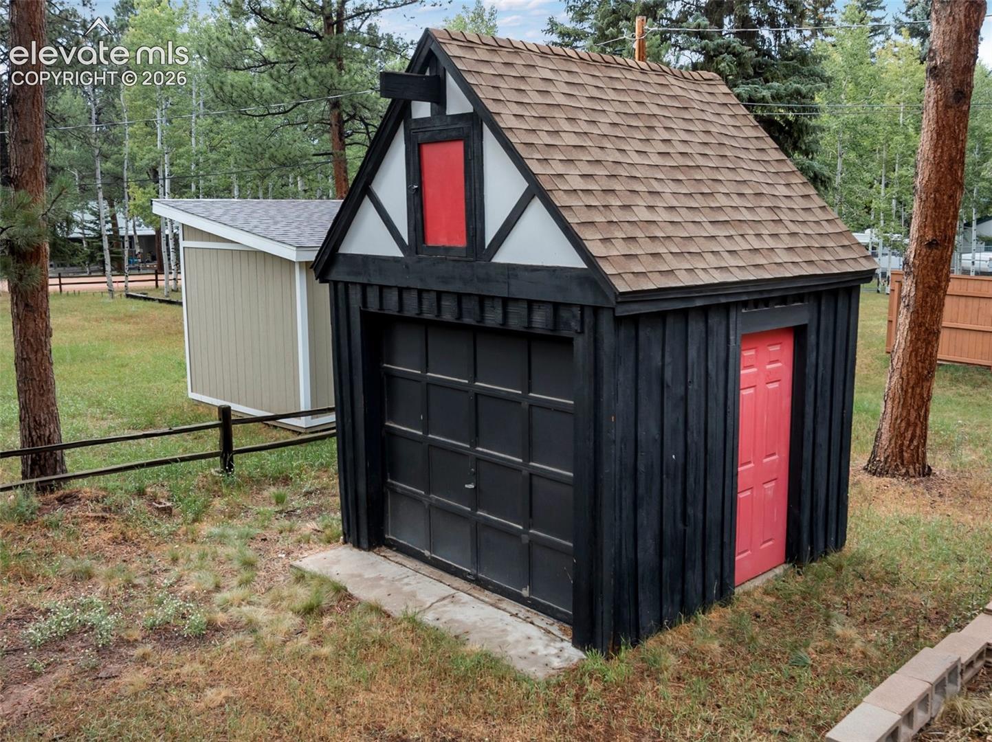 Two distinct outbuildings, one featuring a vertical wood plank exterior with a red entry door and an overhead garage door, and a second structure with horizontal siding and a pitched roof