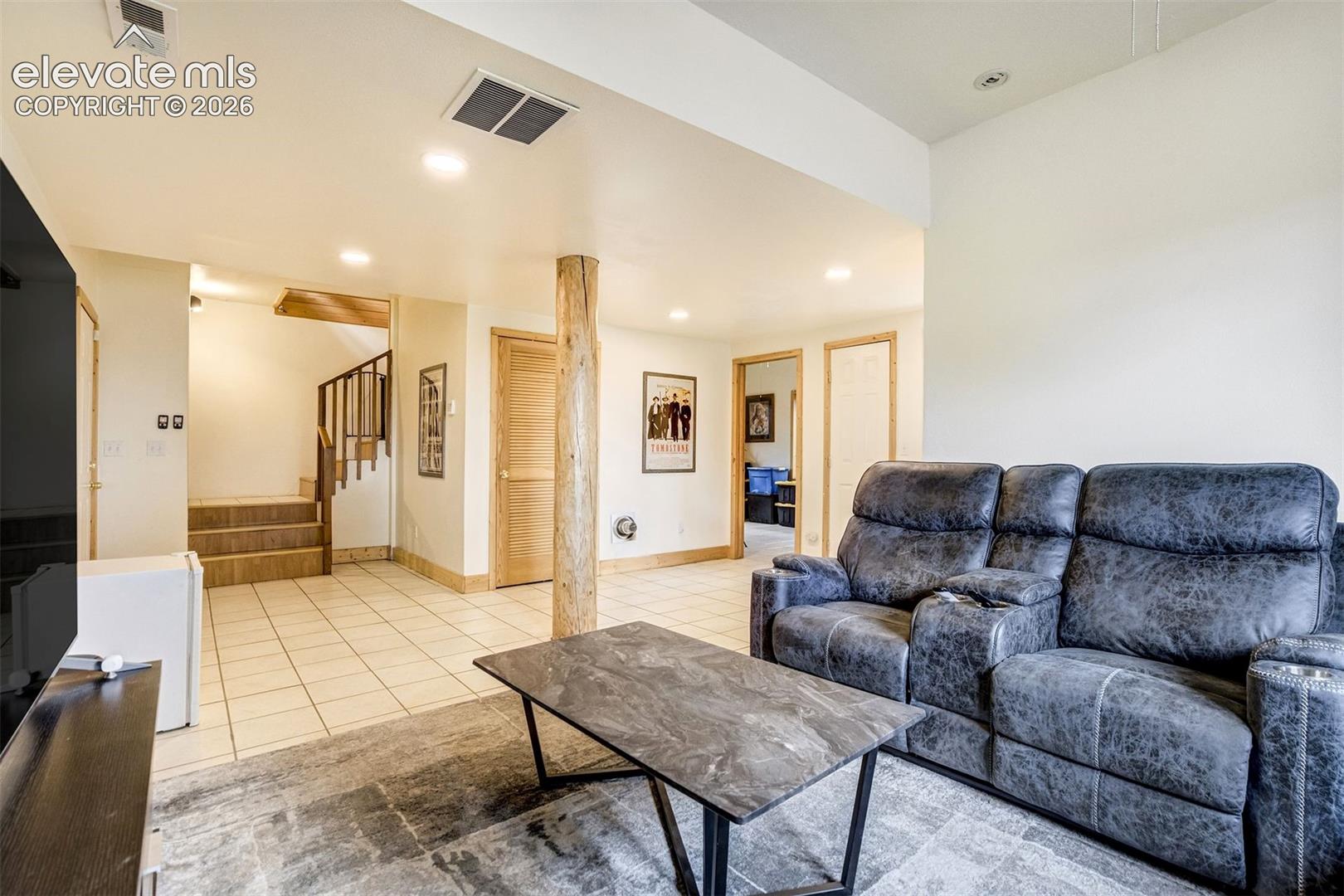 Spacious room featuring light-toned tile flooring, recessed lighting, a natural wood support pillar, a wood-finish staircase with a dark railing, and multiple paneled wood doors