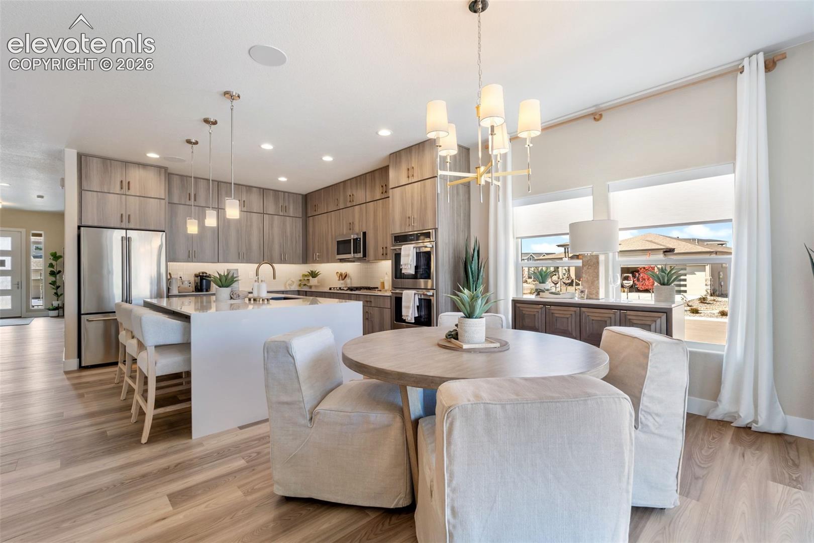 Open-concept kitchen and dining area featuring custom wood cabinetry, stainless steel appliances, quartz countertops, a center island with seating, wood-finish flooring throughout, and a modern pendant light fixture suspended above the dining table