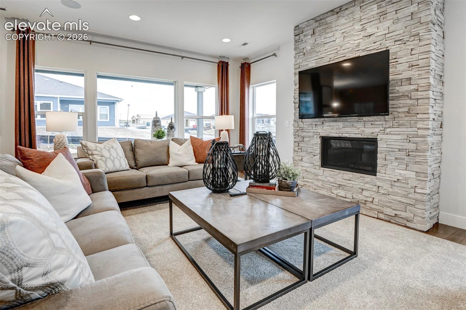 Expansive wall of windows, recessed ceiling lighting, stone-clad fireplace with built-in hearth, and warm wood-finish flooring