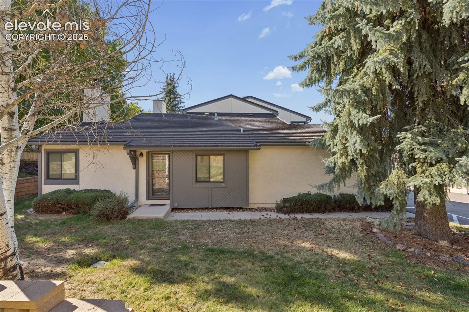 Exterior featuring a stucco facade, dark-tone shingle roofing, concrete walkway, and mature evergreen and birch trees