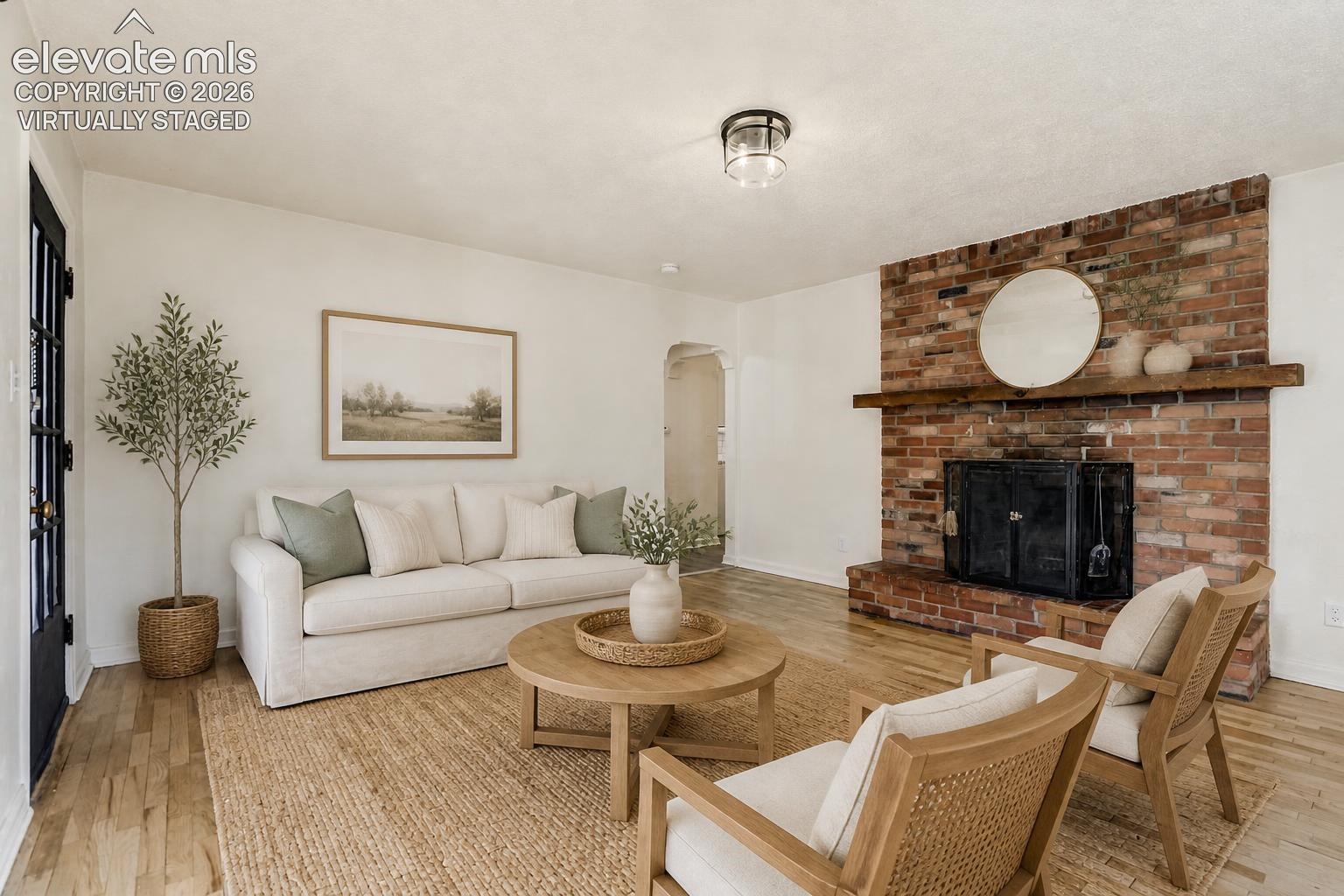 Virtually Staged.  Living area featuring wood-finish flooring, a full-height exposed brick fireplace, a prominent timber mantel, and a multi-pane glass entry door