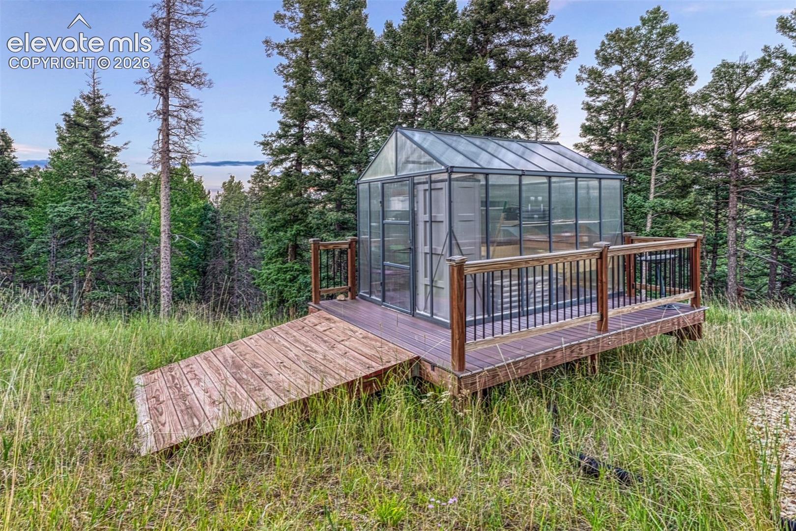 Glass-paneled greenhouse with a pitched roof, set on a raised wood-finish deck with dark railings