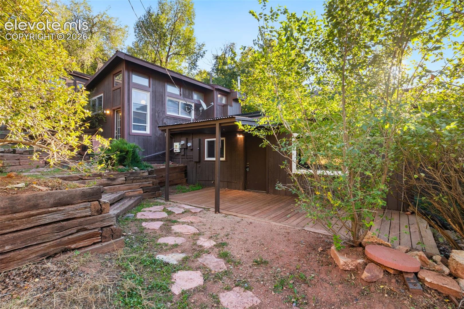 Wood-sided exterior featuring tiered timber retaining walls, a stone paver path, and a covered wood deck
