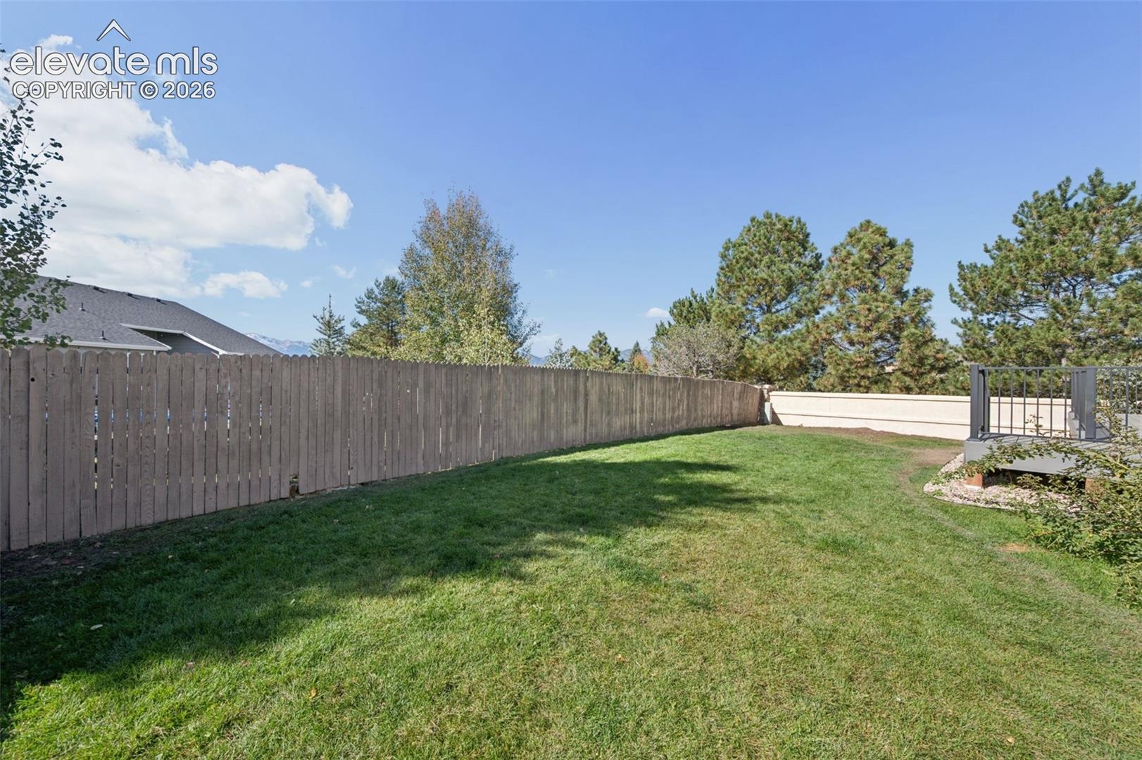 Fenced-in backyard featuring a grass lawn, mature trees, and an elevated composite deck with metal railing