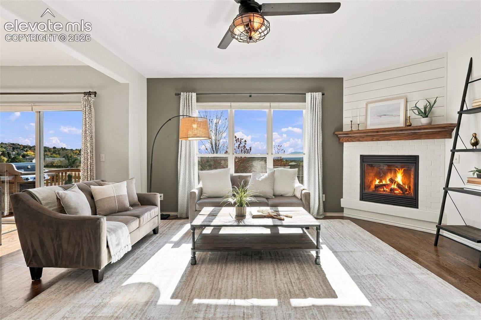 Living area featuring wood-finish flooring, a fireplace with a white painted brick surround and wood mantel, horizontal wall paneling, and a ceiling fan with integrated lighting