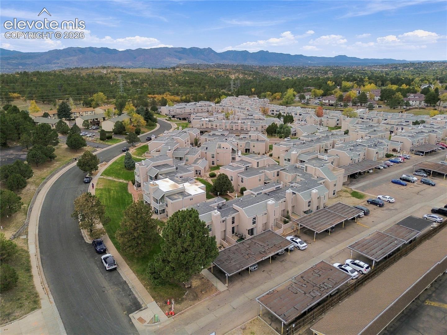 Aerial perspective of a multi-unit residential complex featuring stucco exteriors, shingled roofing, designated carport structures, landscaped grounds, and a backdrop of mountain terrain