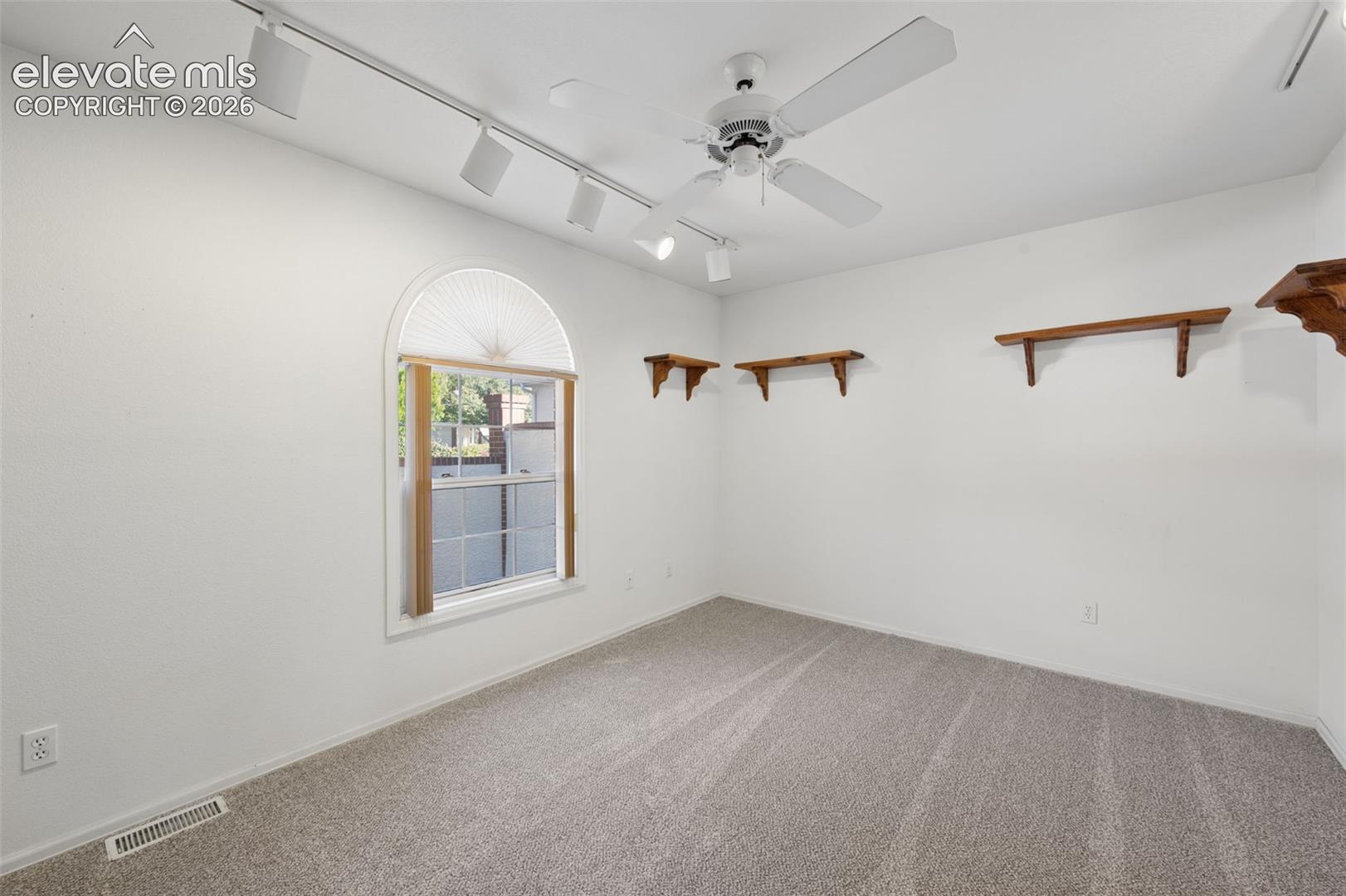 Arched window with casing, ceiling-mounted track lighting, multi-blade ceiling fan, wall-mounted wood-finish shelving, and wall-to-wall carpeting