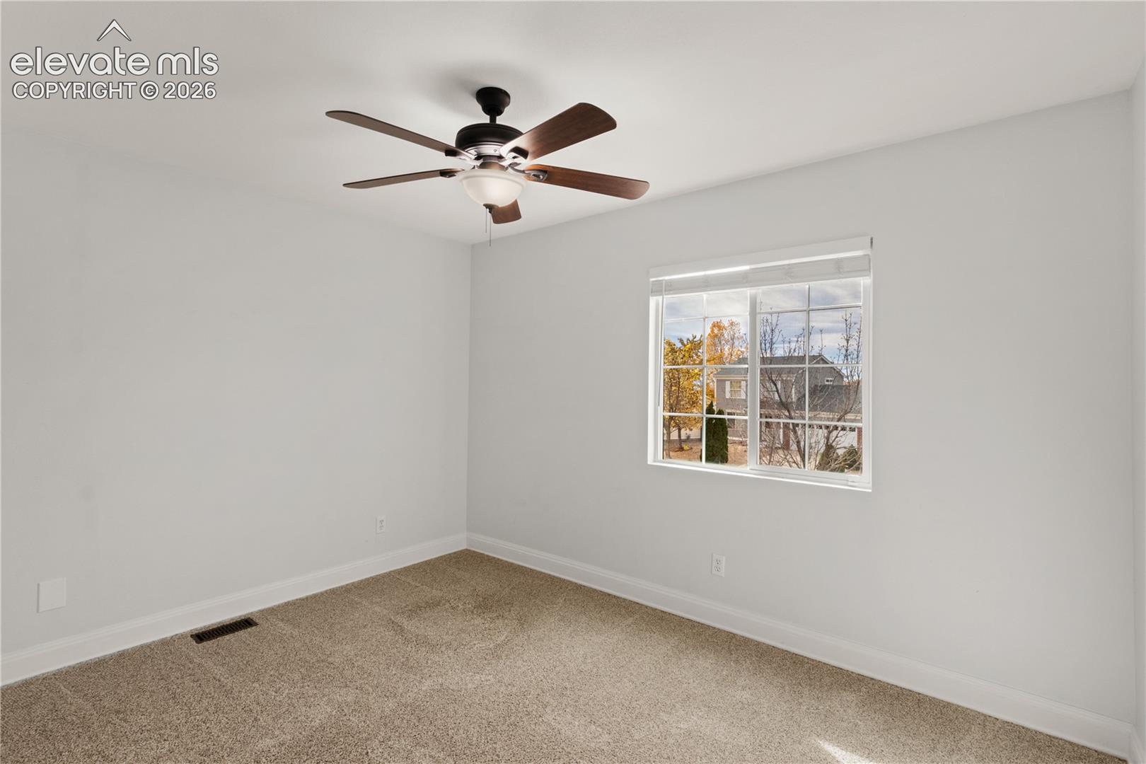Neutral-toned wall paint, wall-to-wall carpeting, and a multi-blade ceiling fan with a center light fixture, illuminated by a single-pane window with interior mullions