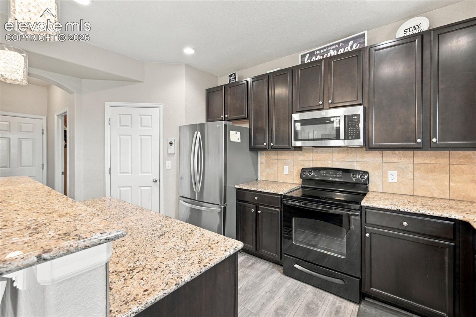 Kitchen featuring dark-finish cabinetry, stone-patterned countertops, a tile backsplash, stainless steel appliances, and wood-finish flooring