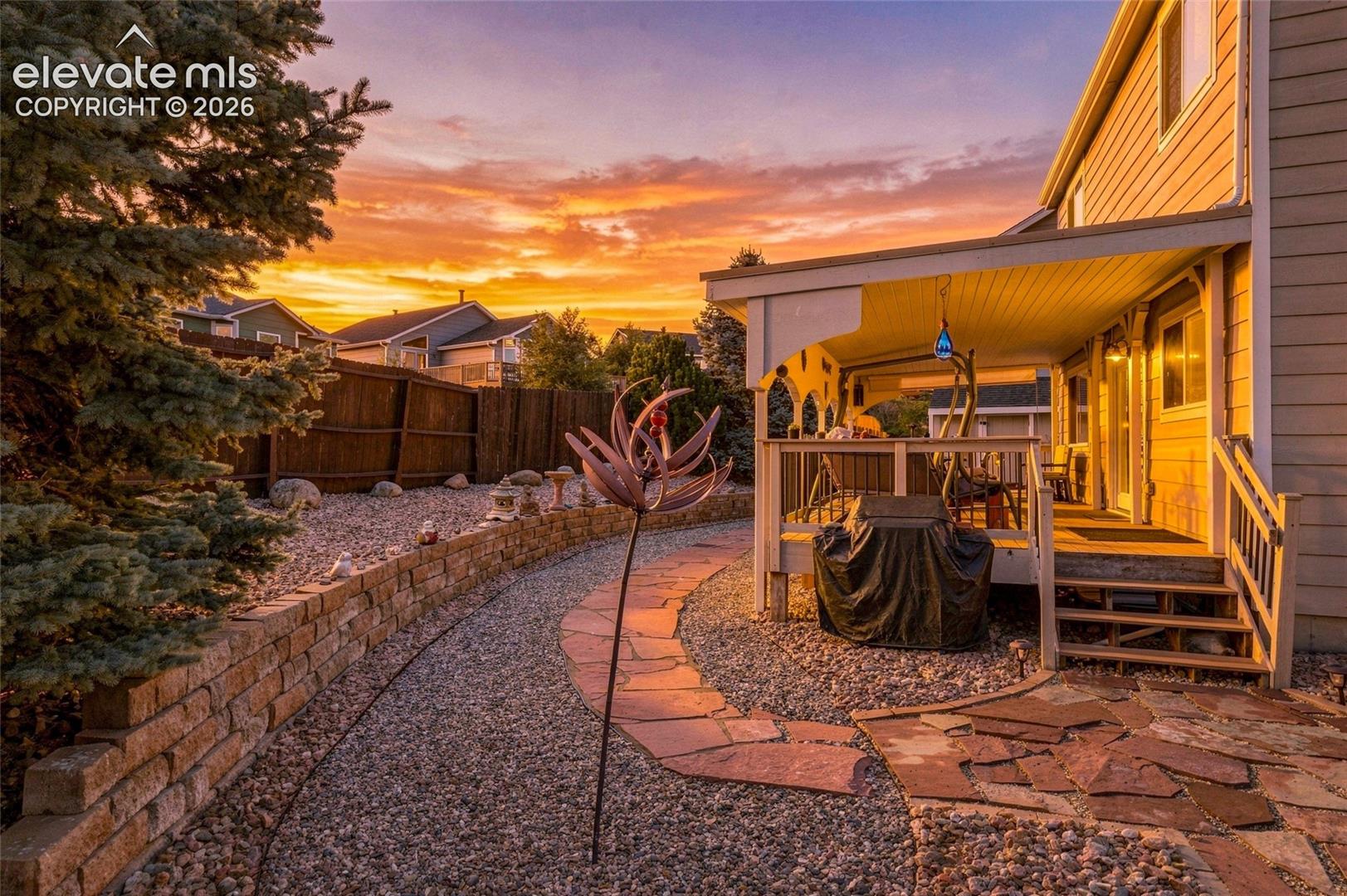 Covered patio with a wood-finish deck and railings, stone paver pathway, gravel landscaping, and a brick retaining wall