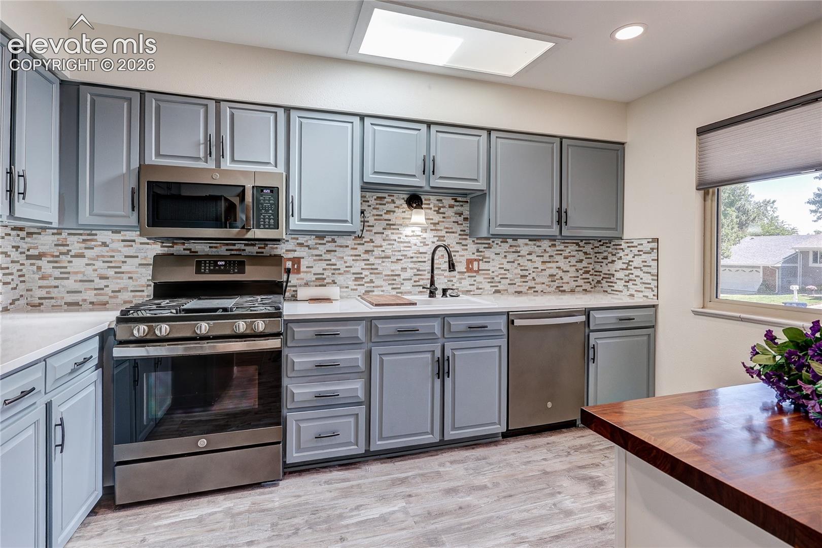 Kitchen featuring shaker-style cabinetry, stainless steel appliances, multi-toned tile backsplash, solid surface countertops, and wood-finish flooring