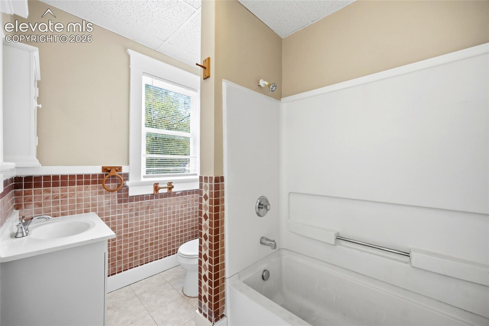 Bathroom featuring a white pedestal sink, a composite tub and shower surround, decorative square-tile wall accents, and a window with wood trim