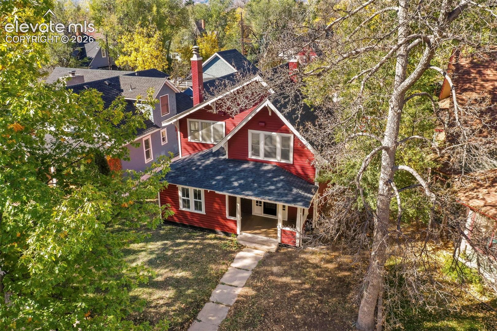Two-story exterior featuring red horizontal siding, a covered front porch, dark shingled roofing, a prominent red chimney, and mature trees