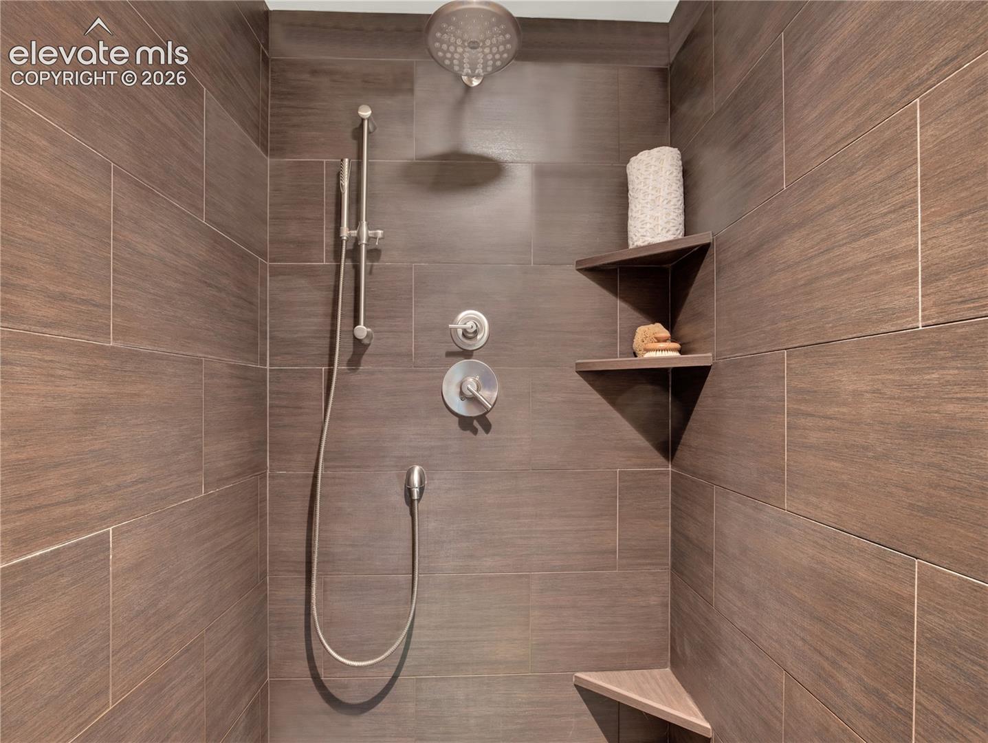 Shower stall featuring wood-finish tile wall surround, brushed nickel rainfall shower head, integrated handheld sprayer, and built-in corner shelving
