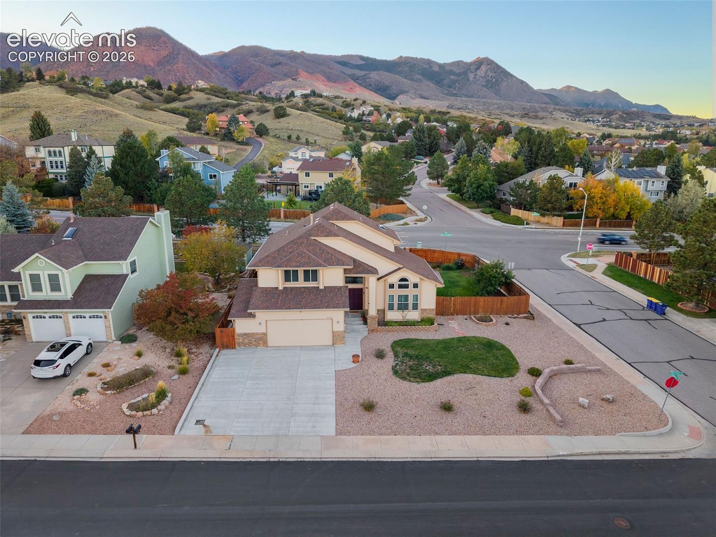 Two-story residence featuring a multi-gabled shingle roof, stone-veneer accents, a multi-car concrete driveway, and xeriscaped front grounds with a boulder retaining wall, set against a mountainous backdrop