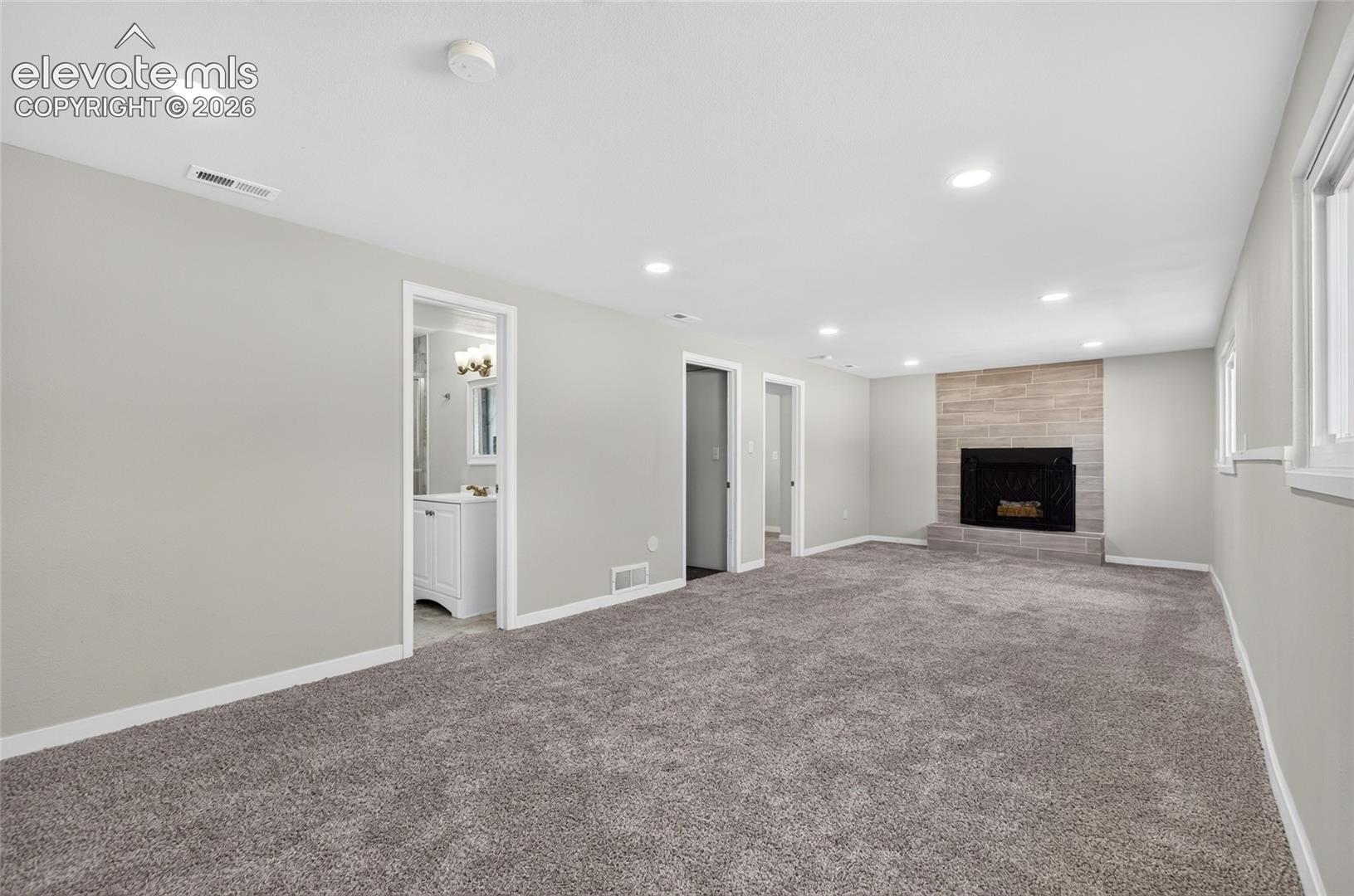Spacious room with plush gray carpeting, recessed ceiling lighting, a stone-faced fireplace with a black firebox, and multiple windows