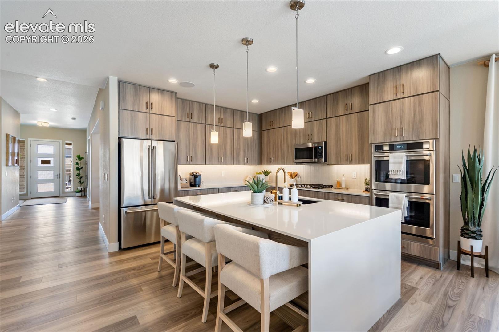 Kitchen featuring full-height wood-finish cabinetry, a waterfall-edge quartz island, stainless steel built-in double ovens, recessed lighting, and pendant fixtures