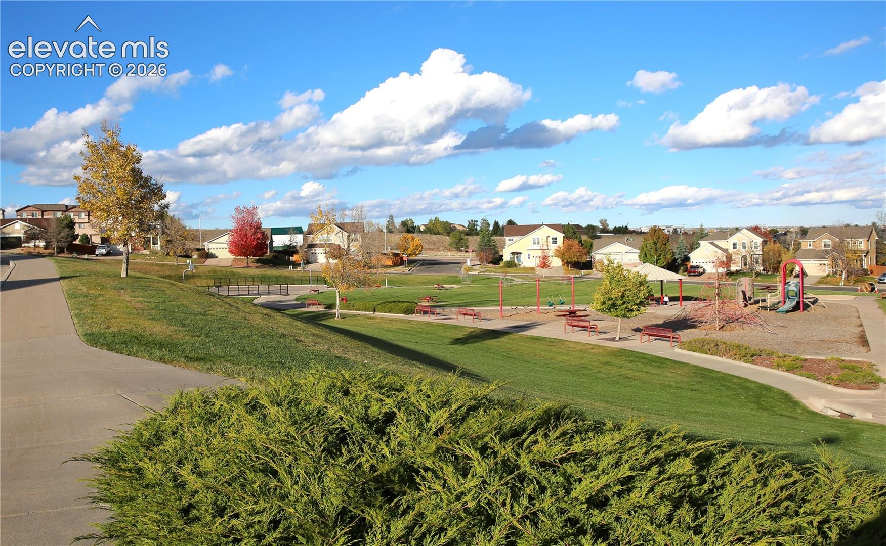 Open park area featuring a paved pedestrian walkway, manicured grass lawns, playground equipment, and mature trees
