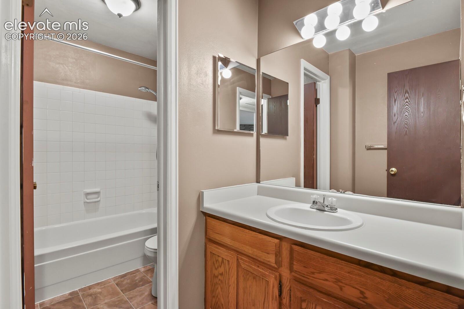 Bathroom featuring a wooden vanity with single sink, a full-width vanity mirror, a multi-bulb light fixture, and a tile-surround bathtub, newer linoleum flooring