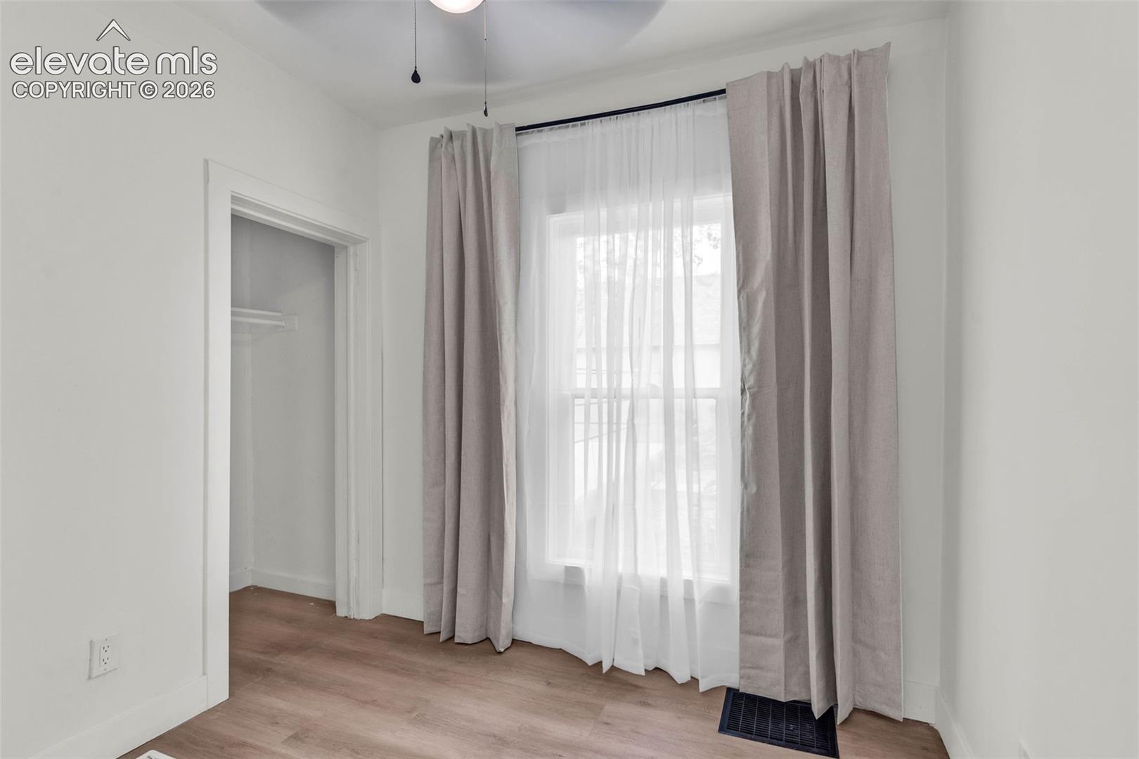 Wood-finish flooring, tall window with neutral-toned treatments, and ceiling fan with integrated lighting
