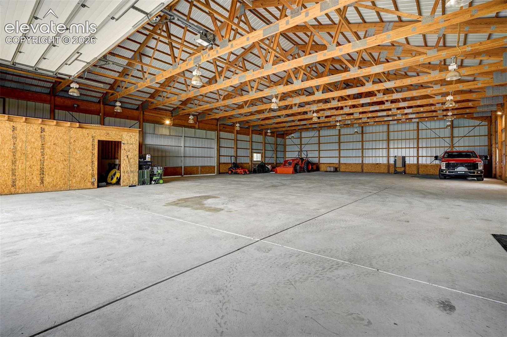 Expansive concrete floor, exposed wood beam ceiling, metal roof, multiple overhead lights, and a built-in enclosed storage area