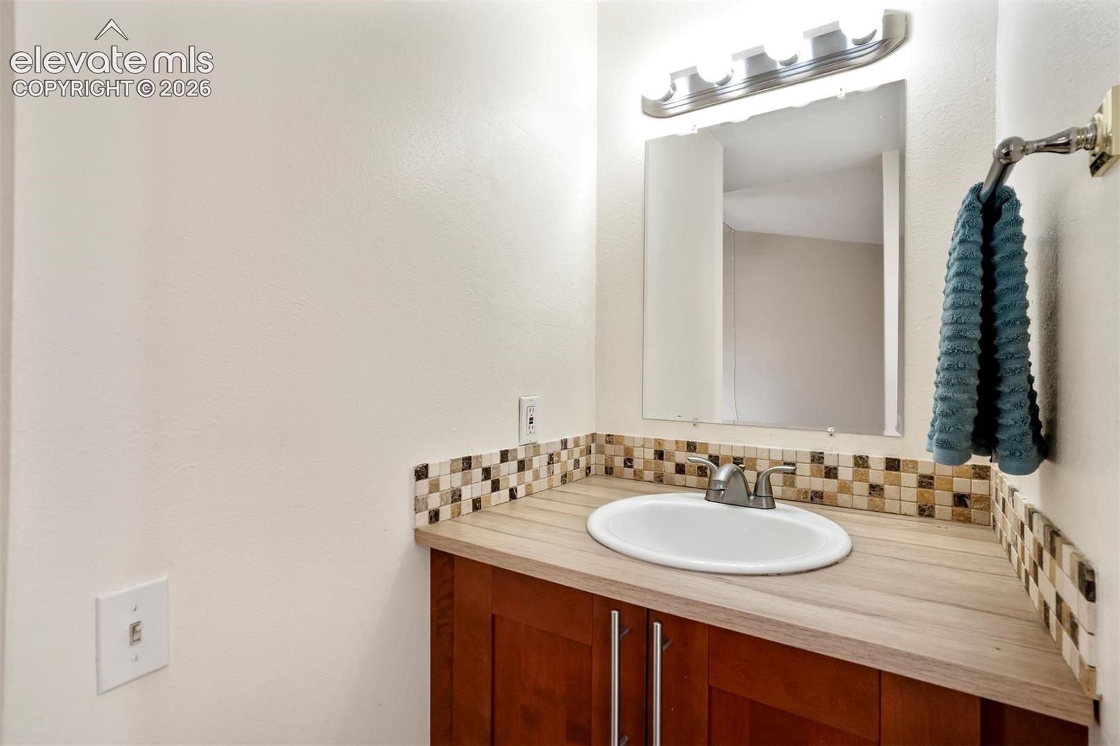 Bathroom vanity featuring wood-finish cabinetry, integrated countertop with a drop-in sink, multi-tone mosaic tile backsplash, and wall-mounted vanity lighting