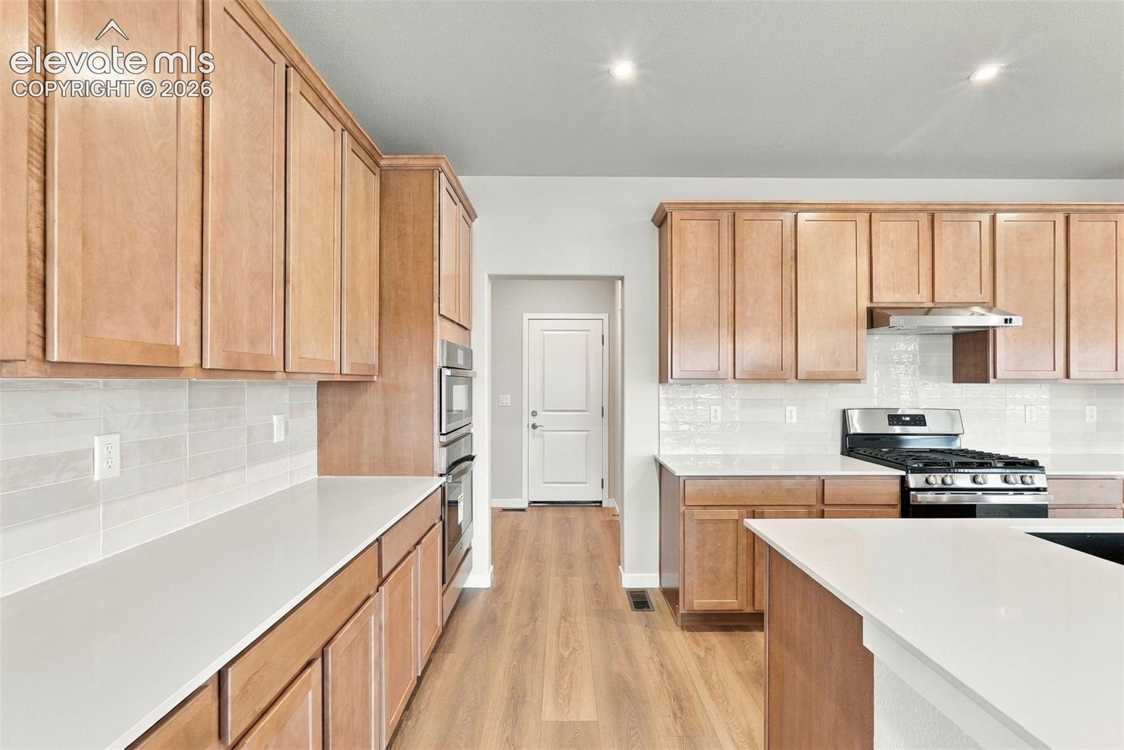 Kitchen featuring shaker-style cabinetry, solid-surface countertops, subway tile backsplash, stainless steel built-in oven and microwave, and wood-finish flooring