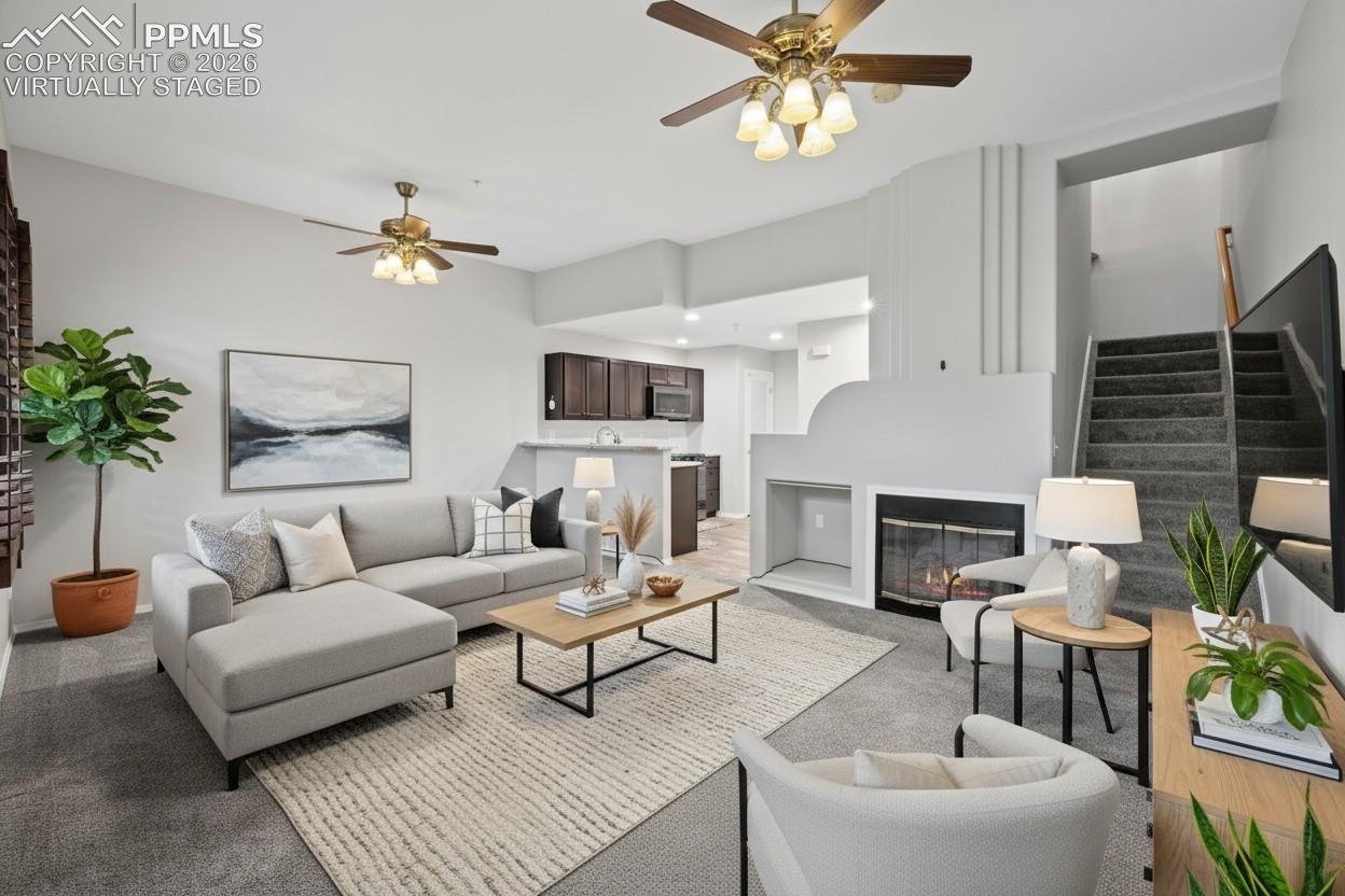 Living room featuring custom window shutters, a ceiling fan, carpet flooring, stairway, and a glass covered fireplace. This photo is virtually staged
.