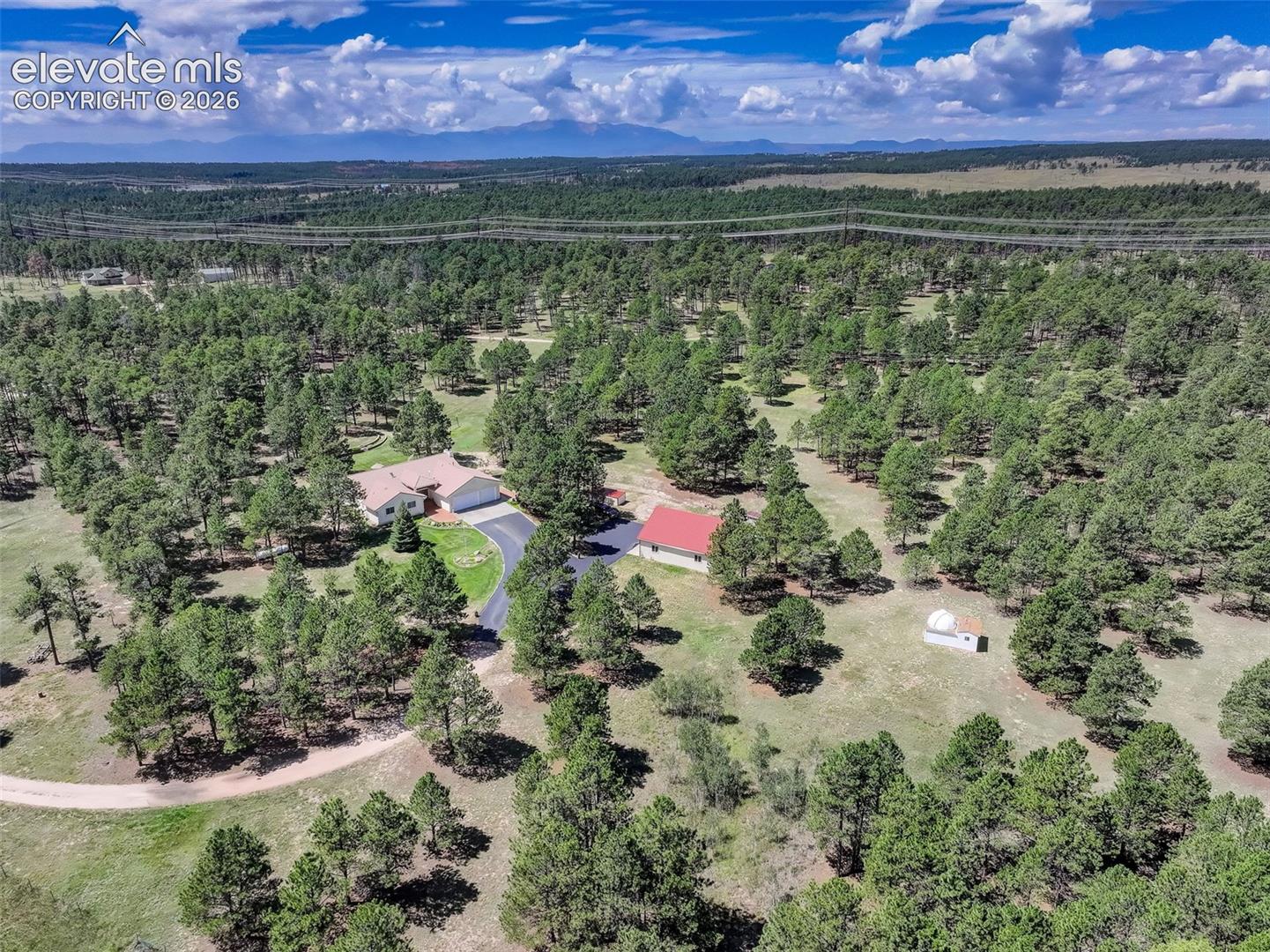 Expansive property featuring a primary residence, RV outbuilding, firewood shed, observatory, a circular driveway, mature evergreen trees, and a distant mountain range.