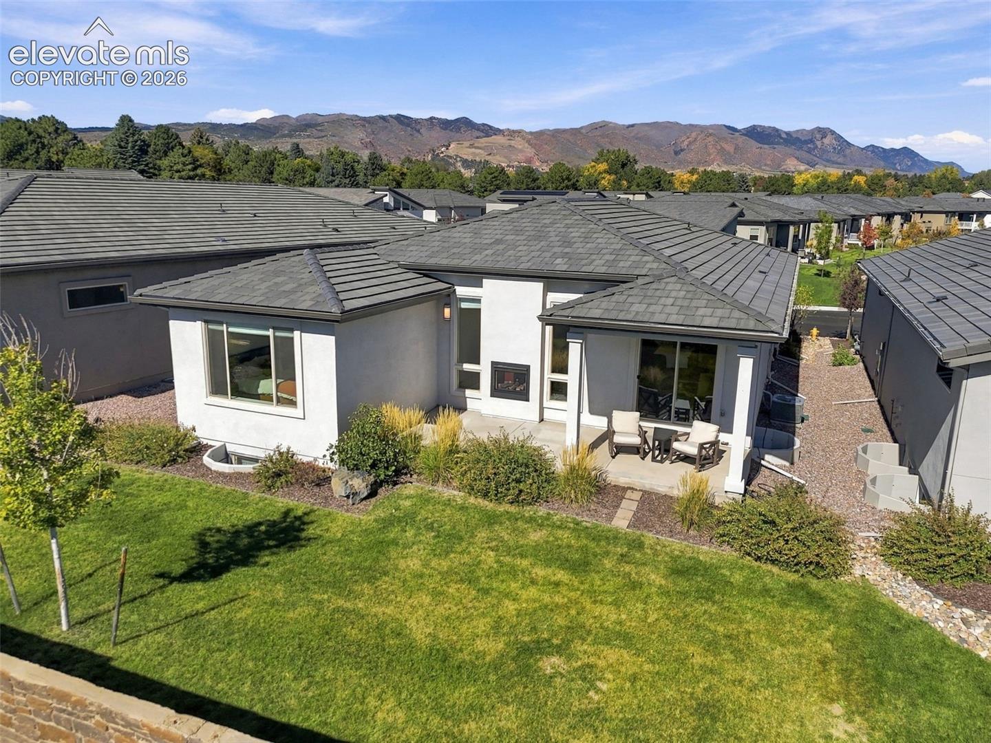 Rear exterior featuring stucco siding, a tiled roof, a built-in outdoor fireplace, a covered concrete patio, and a landscaped lawn against a mountain backdrop