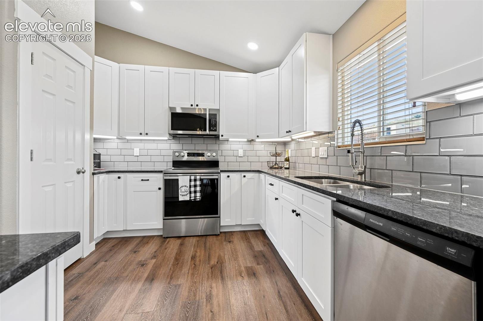Kitchen featuring white shaker-style cabinetry, granite countertops, stainless steel appliances, subway tile backsplash, pantry and wood-finish flooring