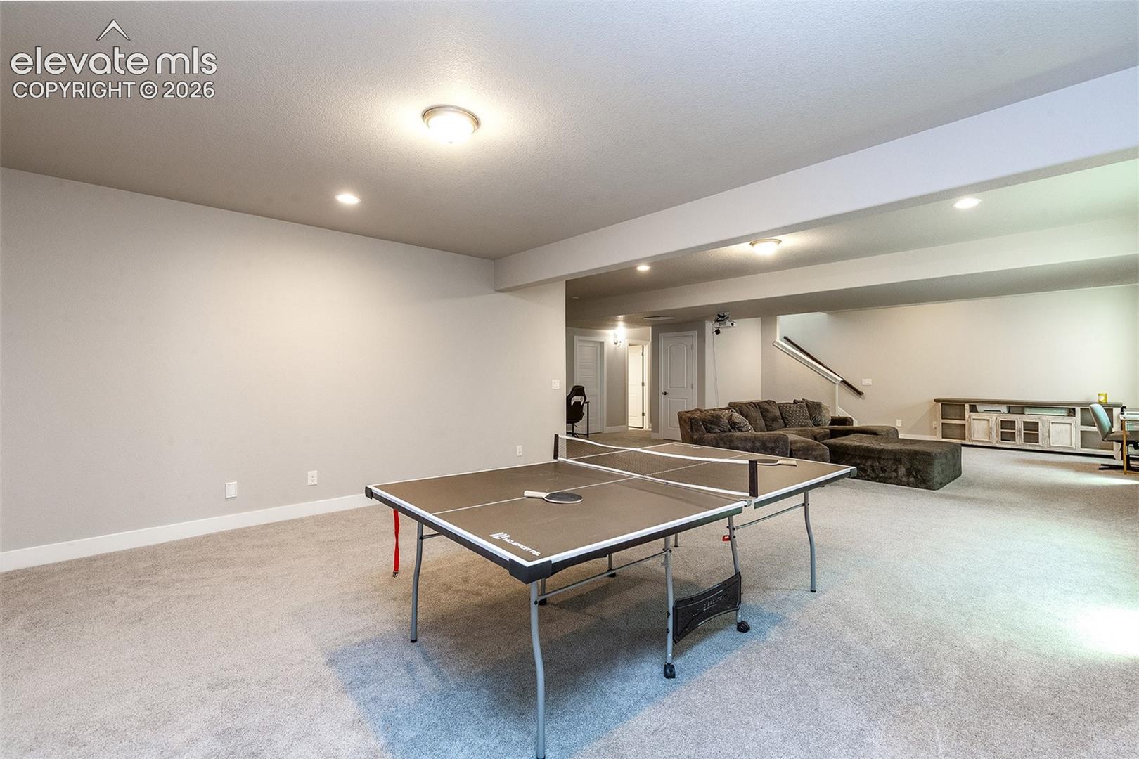 Expansive room with neutral carpet, recessed lighting, and a visible staircase with dark railings
