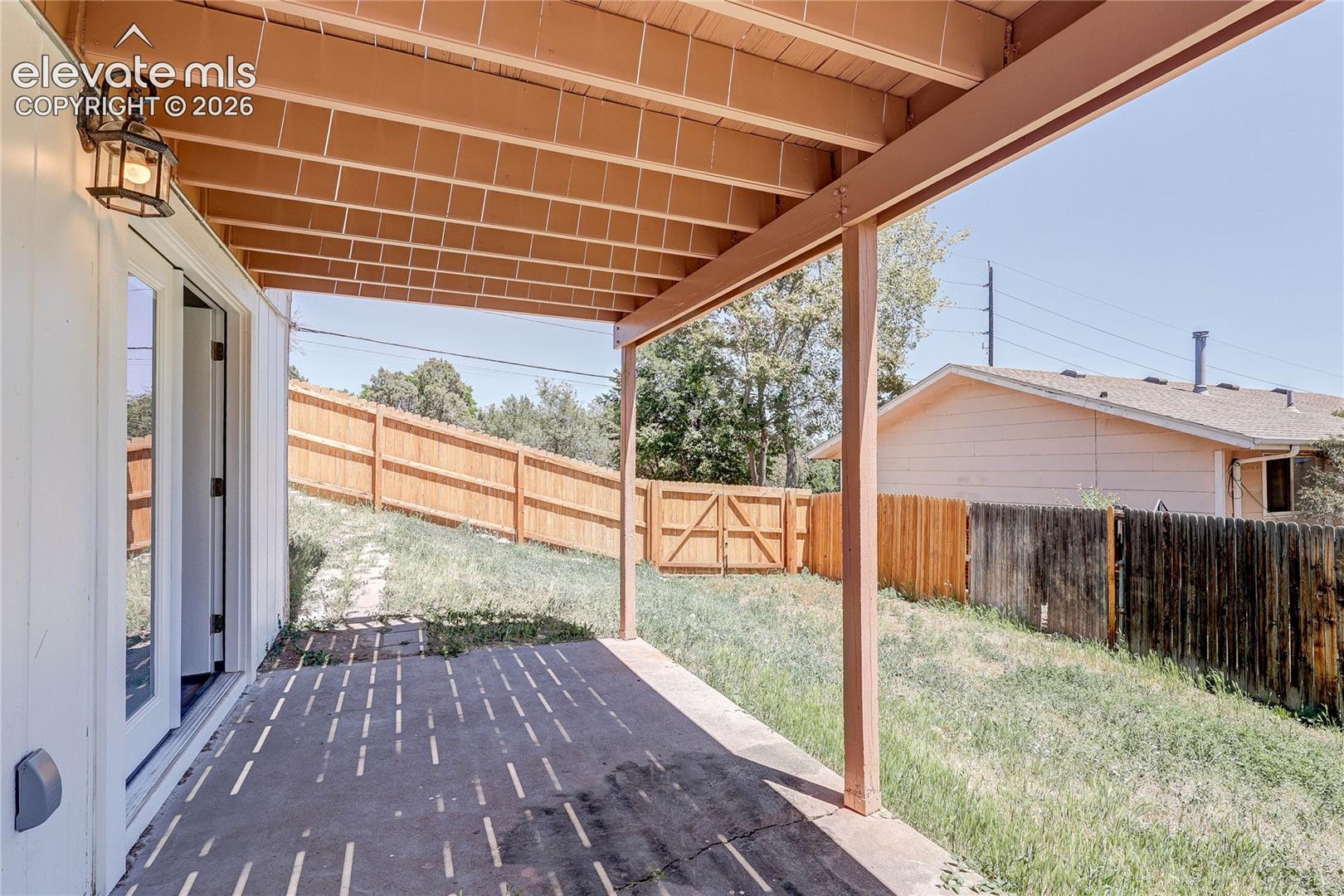 Covered concrete patio featuring an overhead beam structure, exterior wall-mounted lighting, and a fenced yard with a wooden gate