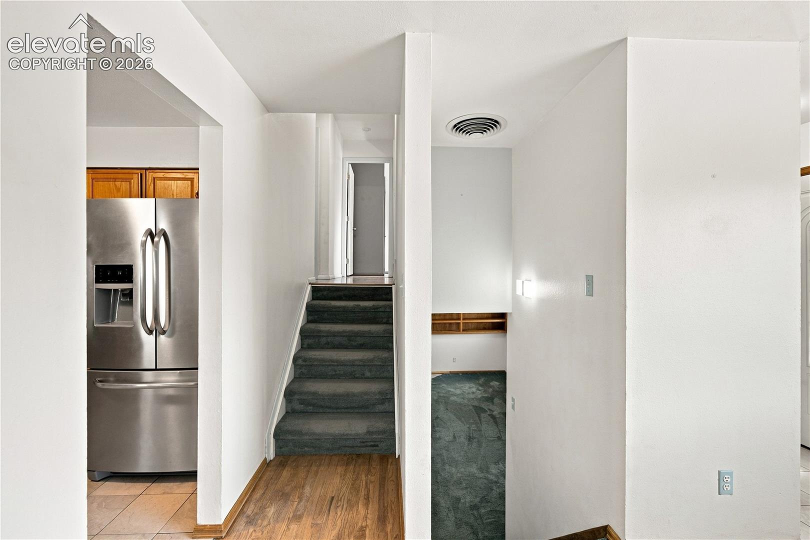 Stainless steel refrigerator, wood-finish flooring, exposed shelving, and carpeted staircase