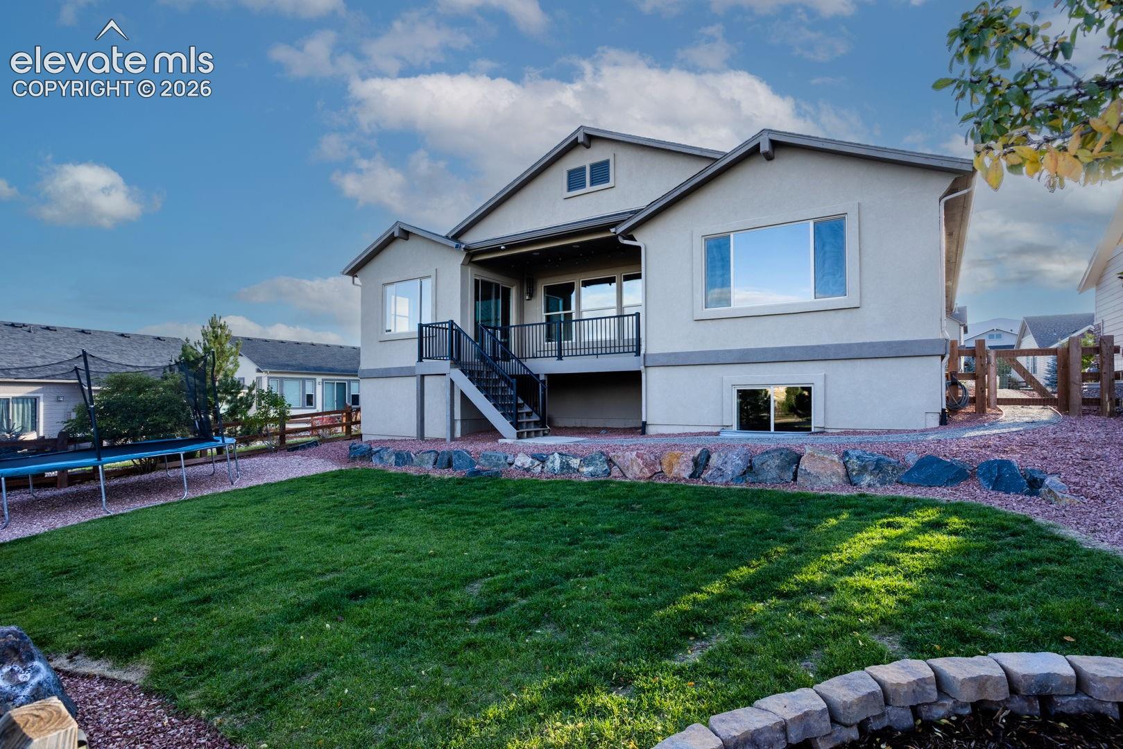 Stucco exterior featuring an elevated deck with metal railing, stone retaining wall, manicured lawn, and perimeter wood fencing