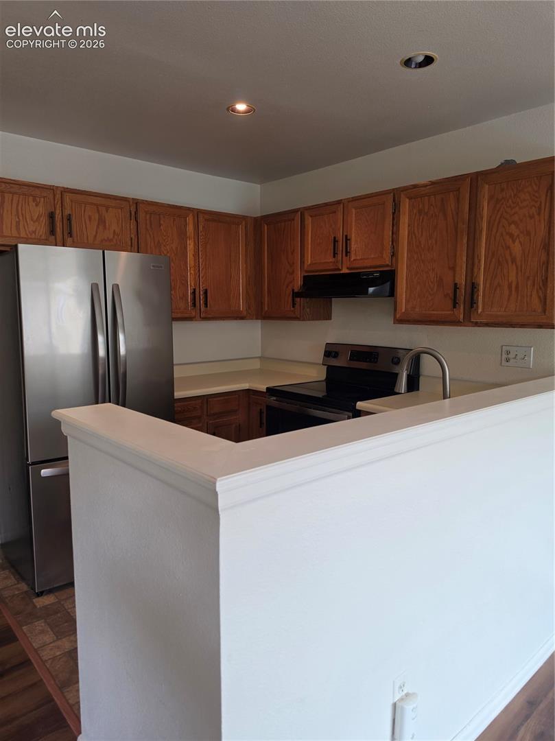 Kitchen featuring wood cabinetry, solid surface countertops, stainless steel refrigerator, black range, and recessed ceiling lighting