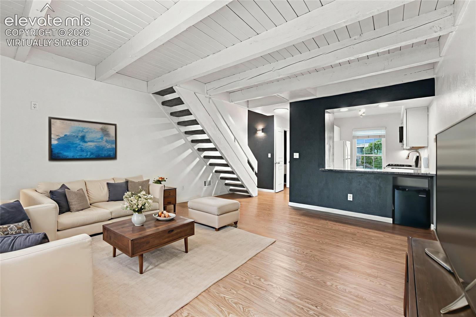 Living area featuring wood-finish flooring, exposed white-painted ceiling beams, a staircase with white metal railings, and a kitchen pass-through window with a dark-toned accent wall
Virtually Staged